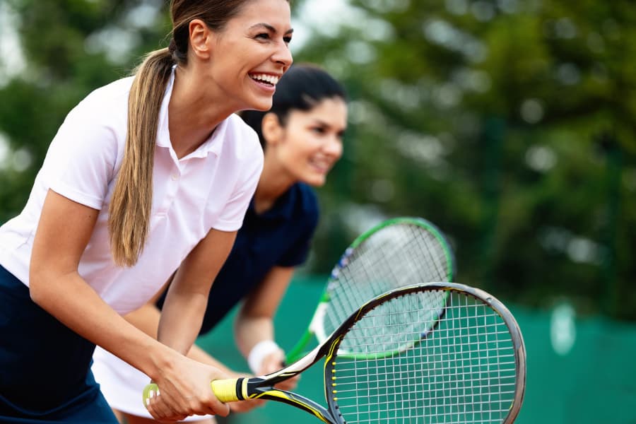 Two tennis doubles players hold racquets and smile as they wait for the ball